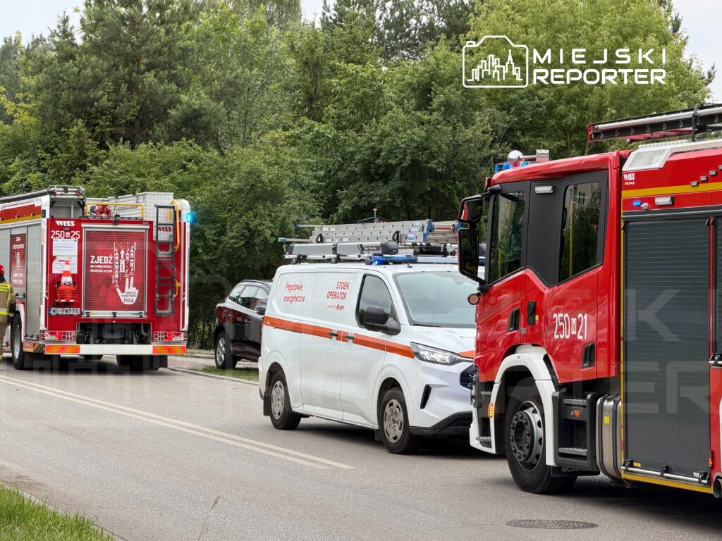 Zespół ratunkowy z wozem strażackim i pojazdem energetycznym zatrzymany na drodze w otoczeniu zieleni.