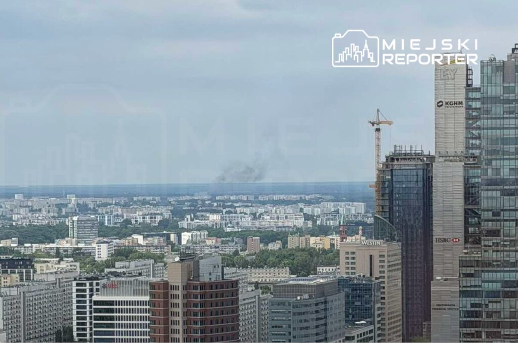 Widok na miejską panoramę z dymem unoszącym się nad horyzontem w tle, w otoczeniu nowoczesnych budynków.