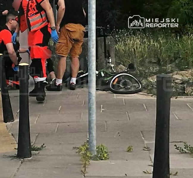 Ratownicy medyczni udzielają pomocy osobie leżącej na chodniku obok roweru, w otoczeniu miejskim.
