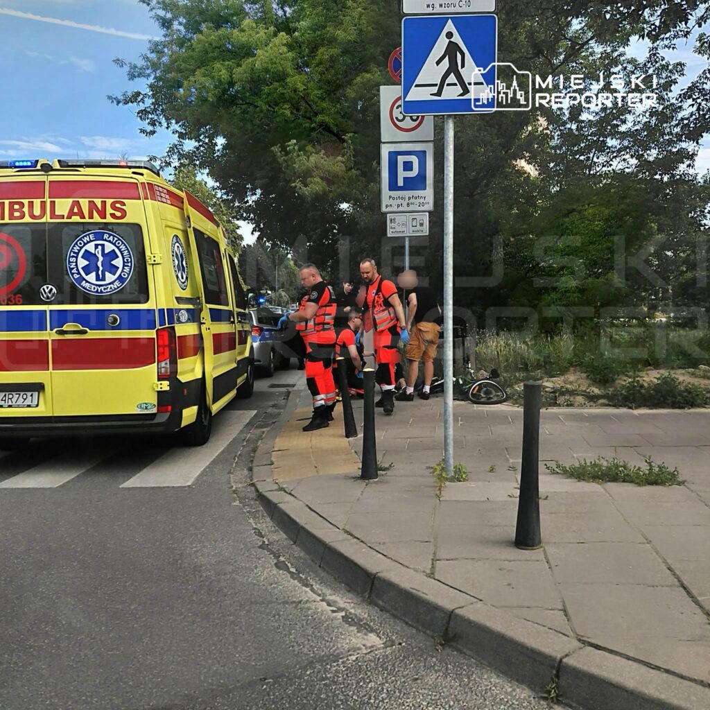 Ratownicy medyczni w pomarańczowych strojach udzielają pomocy poszkodowanemu na chodniku obok karetki.