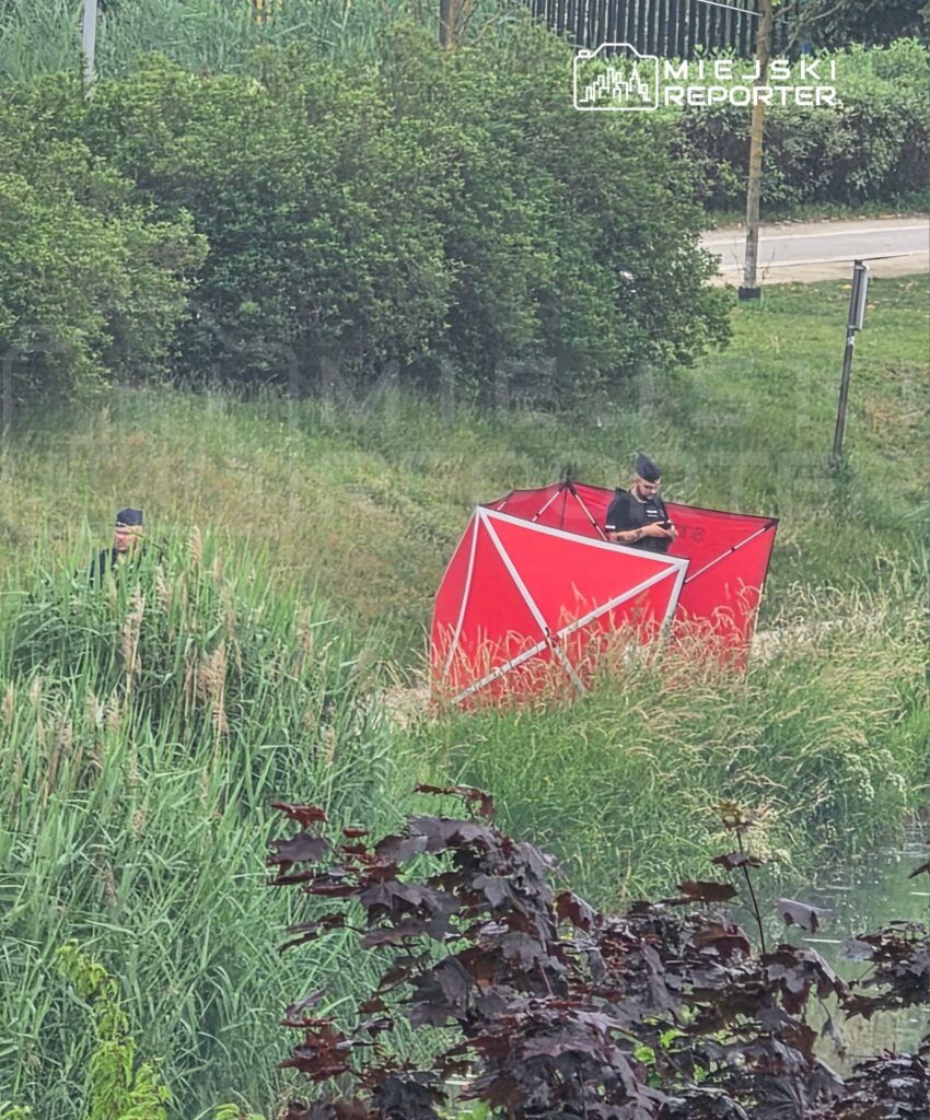 Mężczyzna w mundurze obserwujący teren obok czerwonego namiotu w zaroślach nad wodą.