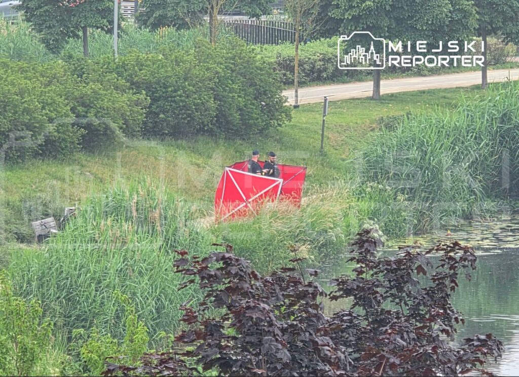 Dwaj mężczyźni w mundurach rozkładają czerwony namiot w zaroślach nad wodą.