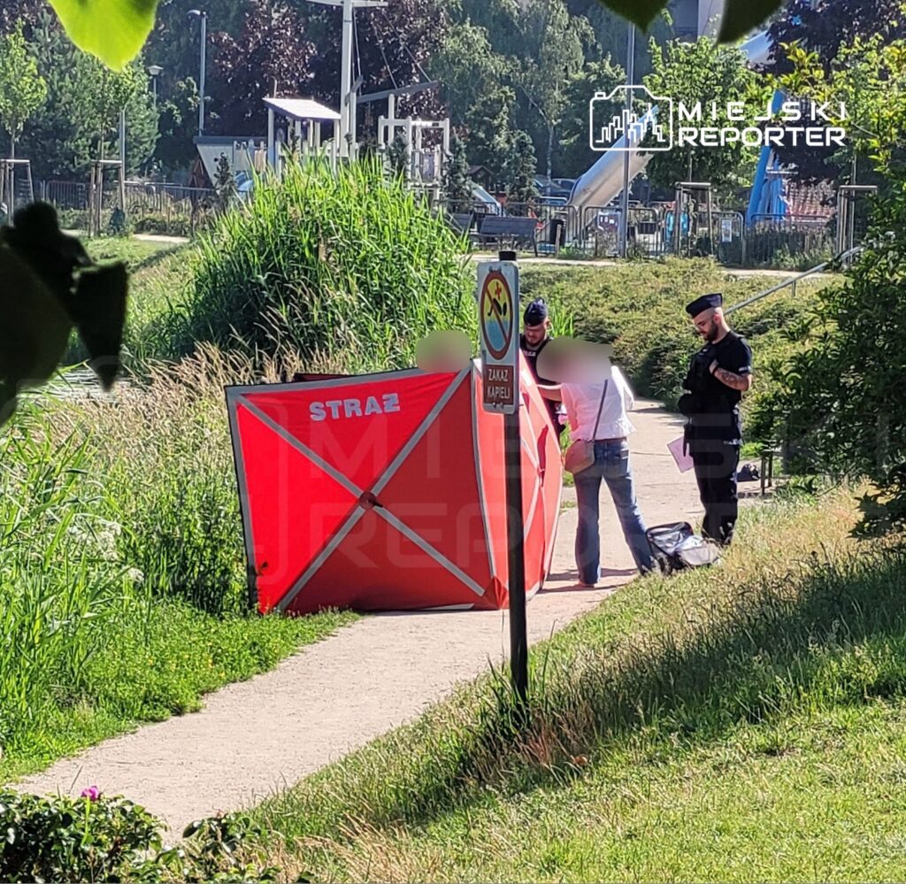 Mężczyzna i kobieta rozmawiają obok namiotu strażackiego na ścieżce w parku, w tle zjeżdżalnia dla dzieci.
