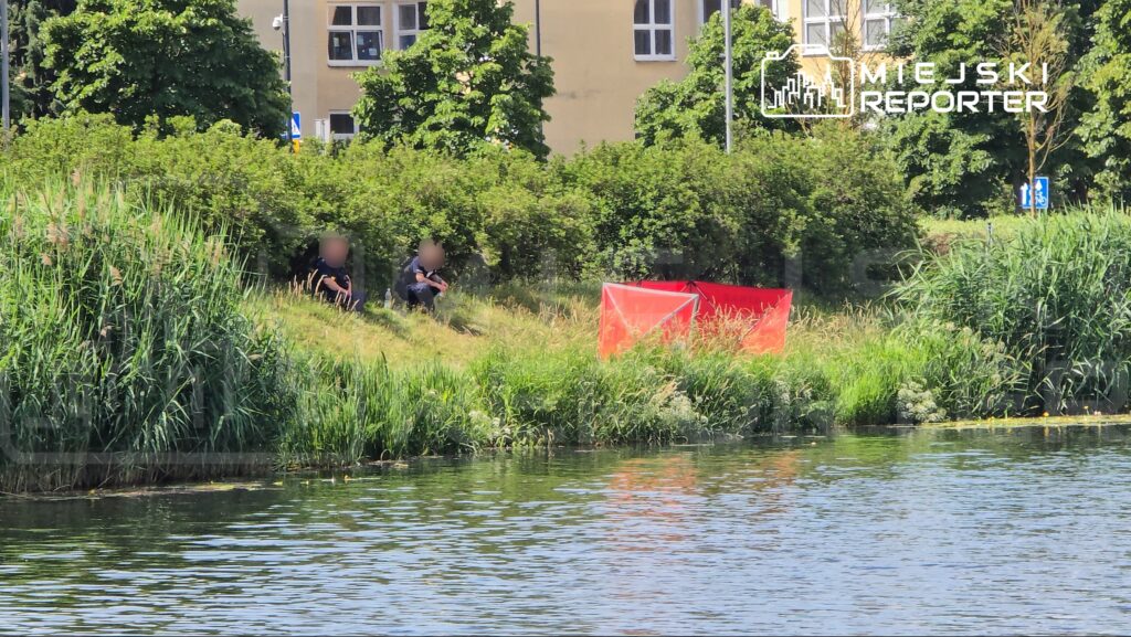 Grupa funkcjonariuszy siedzi na trawie w pobliżu wody, obok czerwonego namiotu w zielonym otoczeniu.