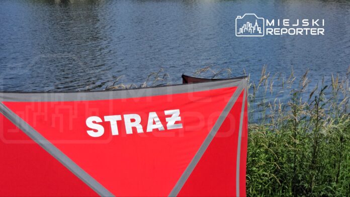 Czerwony namiot z napisem "STRAŻ" ustawiony wzdłuż brzegu wody, otoczony trawą i roślinnością.
