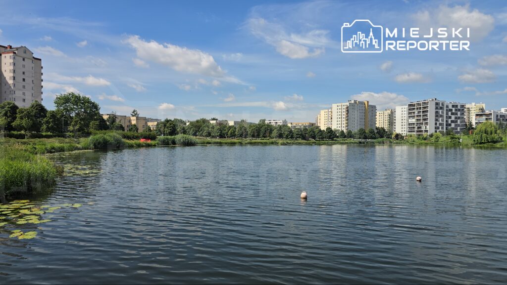 Widok na spokojny zbiornik wodny otoczony zielenią i nowoczesnymi budynkami mieszkalnymi w tle.