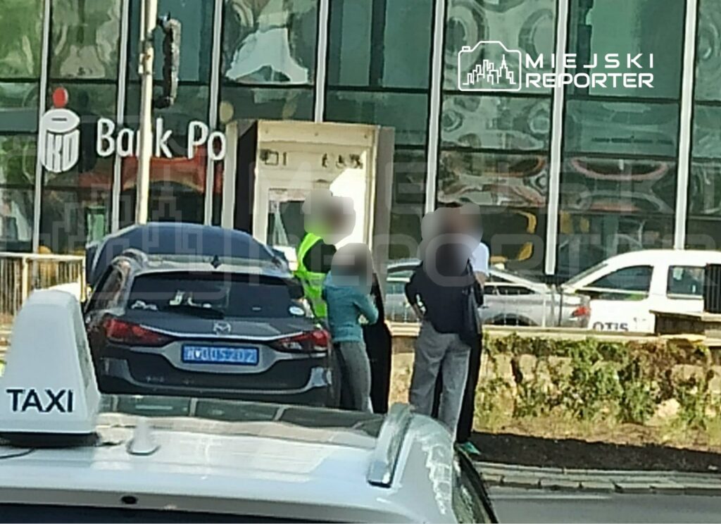 Grupa trzech osób rozmawia obok czarnego samochodu przed nowoczesnym bankiem z przeszkloną fasadą.