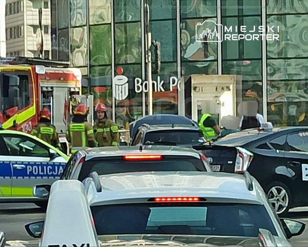 Grupa strażaków w mundurach rozmawia obok wozu strażackiego na ruchliwej ulicy, w tle widoczny bank.