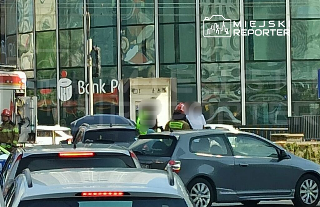 Grupa strażaków i policjantów pracuje przy bankomacie przed nowoczesnym biurowcem, w tle zaparkowane samochody.