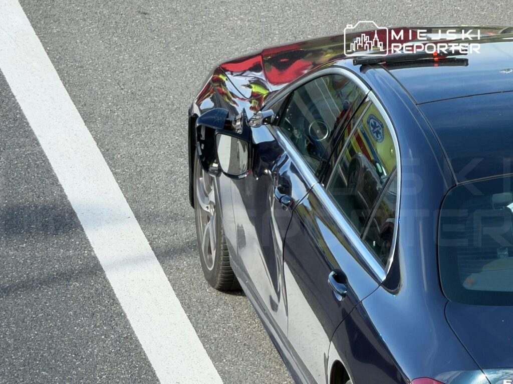 Uszkodzony samochód osobowy z wgniecioną boczną częścią na drodze, obok biała linia oddzielająca pasy.