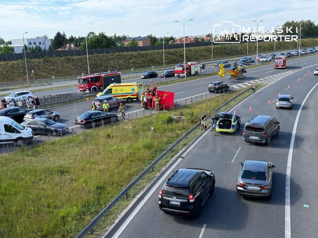 Na drodze szybkiego ruchu zablokowane pojazdy, w tle straż pożarna, karetka i helikopter. Ratownicy udzielają pomocy poszkodowanym.