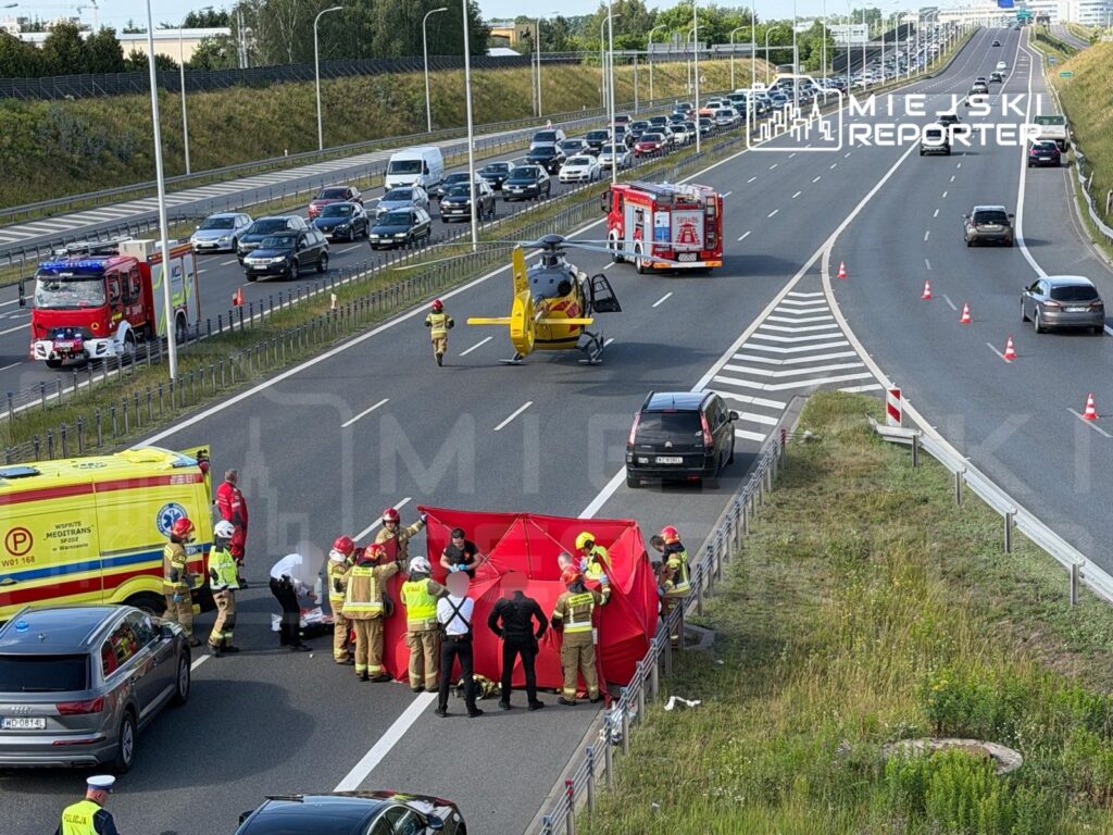 Ratownicy medyczni oraz strażacy udzielają pomocy poszkodowanemu na drodze, obok zjeżdżającego helikoptera.