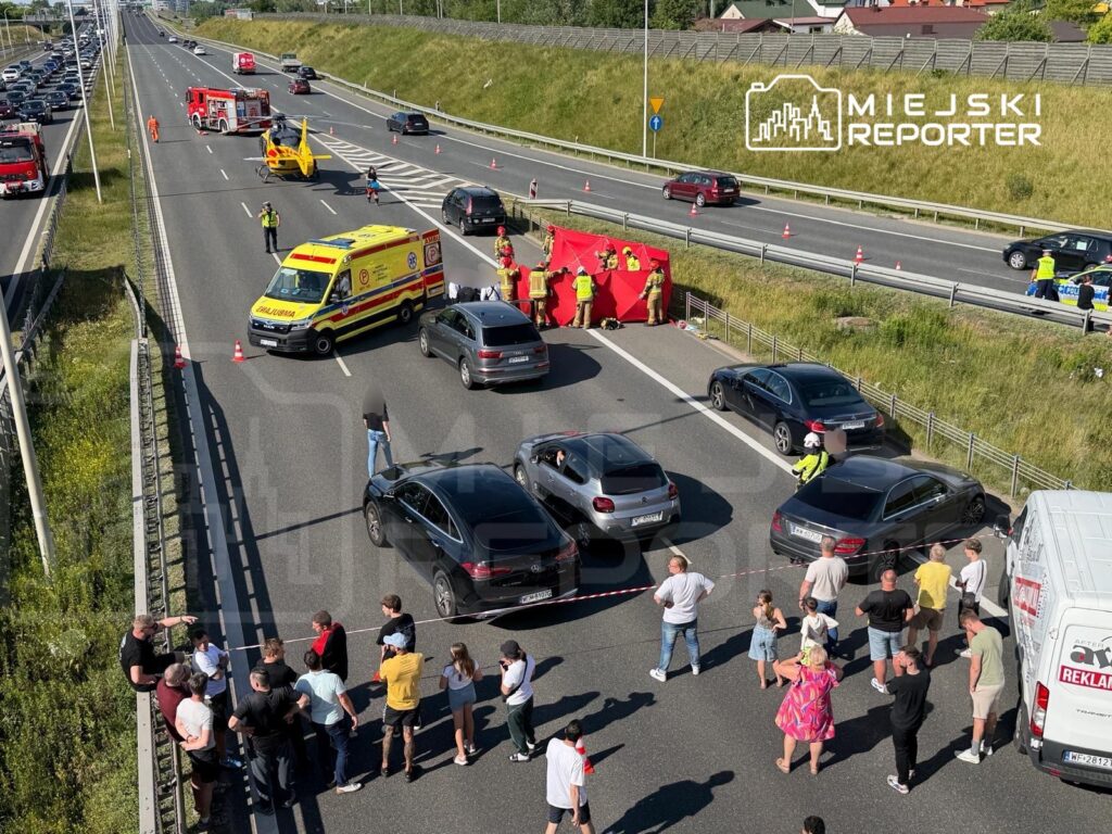 Na drodze ekspresowej zablokowane pojazdy, ratownicy medyczni i strażacy udzielają pomocy poszkodowanym, a tłum gapiów obserwuje sytuację.