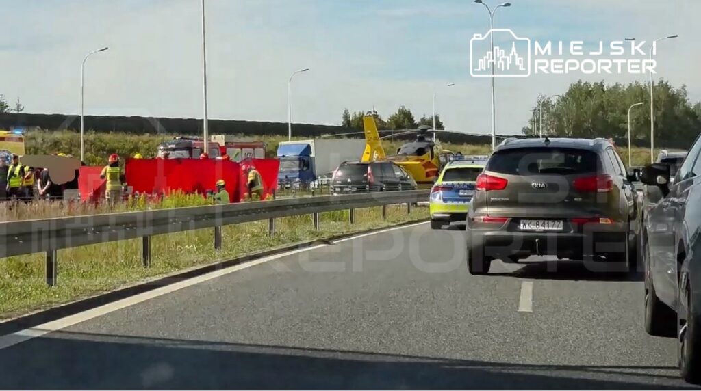 Na drodze widać zespół ratunkowy, w tym strażaków i ratowników, pracujących przy wypadku drogowym. Obok lądował śmigłowiec ratunkowy.