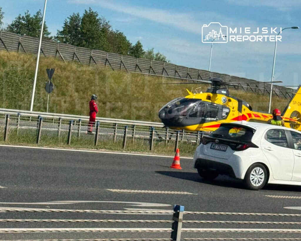 Ratunkowy helikopter lądował na poboczu drogi, obok stoi mężczyzna w czerwonym uniformie.