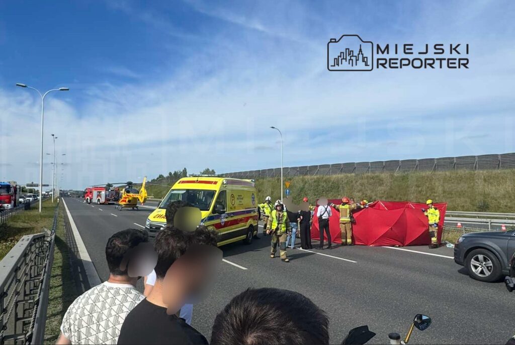 Grupa ratowników i strażaków zabezpiecza miejsce wypadku na drodze, obok stoją karetka i helikopter.