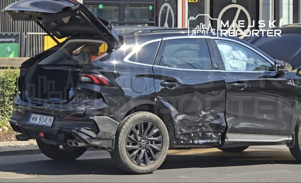 Czarny SUV z uszkodzeniami bocznymi i otwartą klapą bagażnika stoi na ulicy, obok widoczny jest znak drogowy.