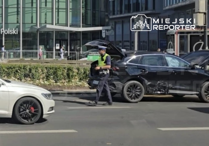 Policjant w zielonej kamizelce kontrolujący czarny samochód na ruchliwej ulicy przed nowoczesnym biurowcem.
