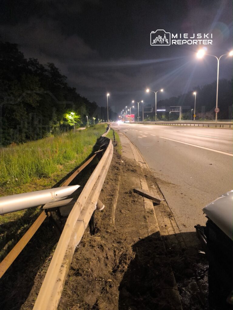 Uszkodzony barierka ochronna przy drodze w nocy, z widocznymi śladami ziemi i trawy obok.