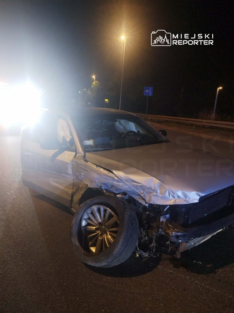 Zniszczony samochód osobowy stoi na drodze, z uszkodzoną przednią częścią i jednym zerwanym kołem, w nocnym otoczeniu.