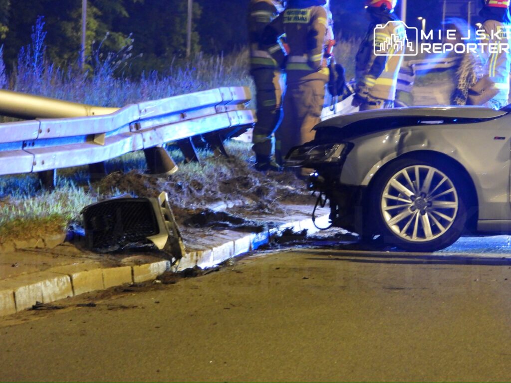 Uszkodzony samochód osobowy stoi na poboczu drogi, a obok niego pracują strażacy w mundurach.