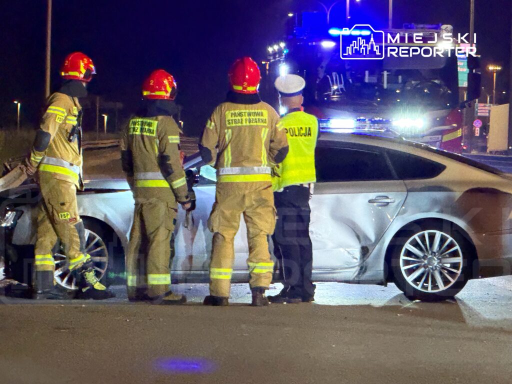 Czwórka strażaków w mundurach i hełmach bada uszkodzone auto w nocy, obok stoi policjant w kamizelce odblaskowej.