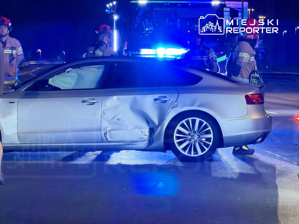 Zniszczony samochód osobowy z wgnieceniem na drzwiach, otoczony przez strażaków w mundurach, podczas nocnej interwencji.