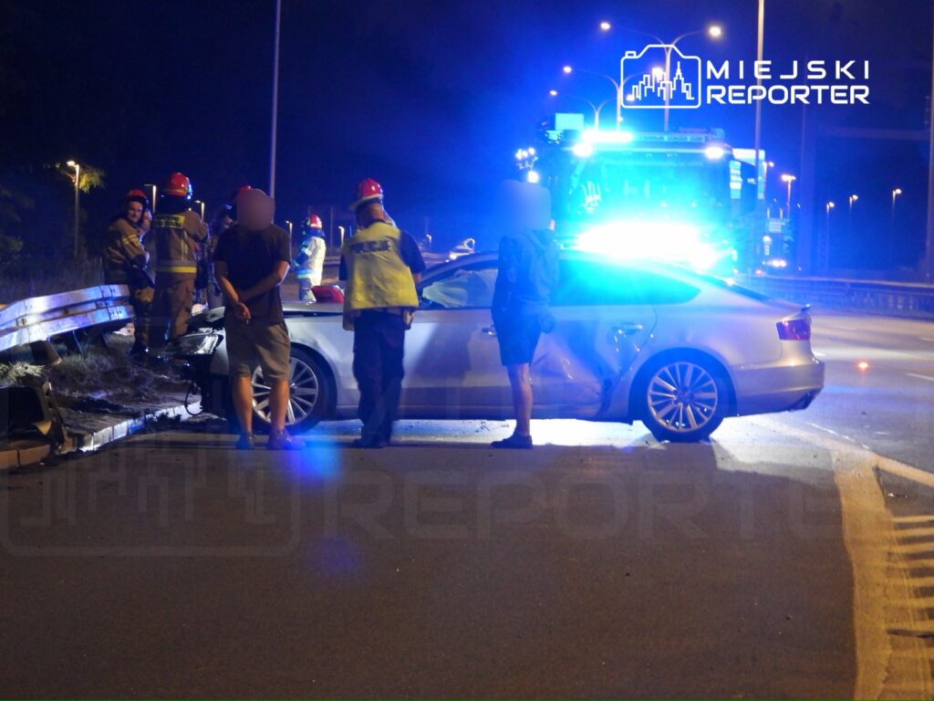Grupa strażaków w kaskach ocenia uszkodzenia samochodu po wypadku na drodze nocą, w tle niebieskie światła radiowozu.