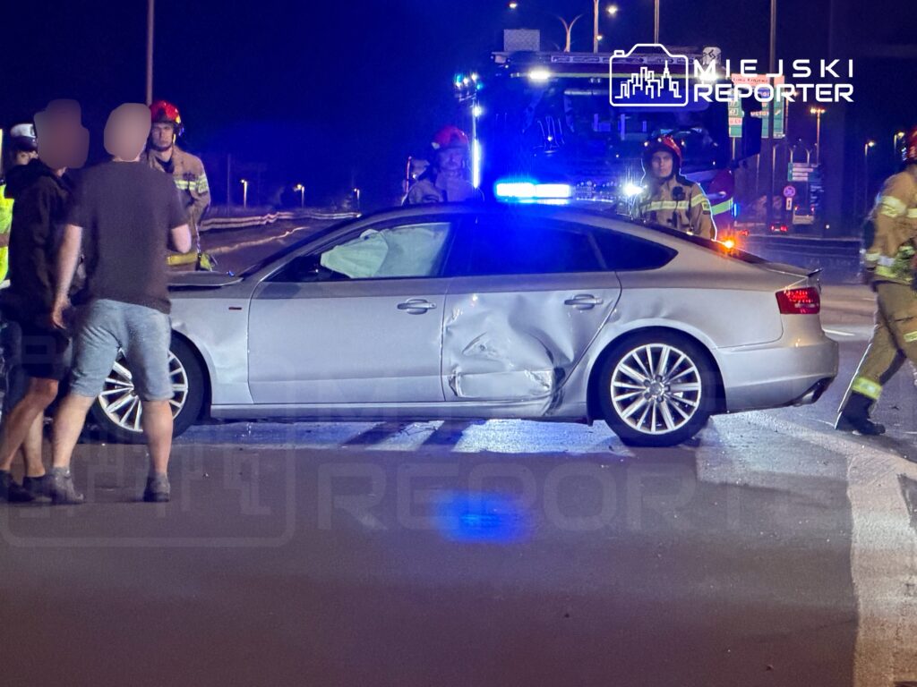 Grupa strażaków i świadków ogląda uszkodzony srebrny samochód na drodze w nocy, obok widać włączone pojazdy ratunkowe.