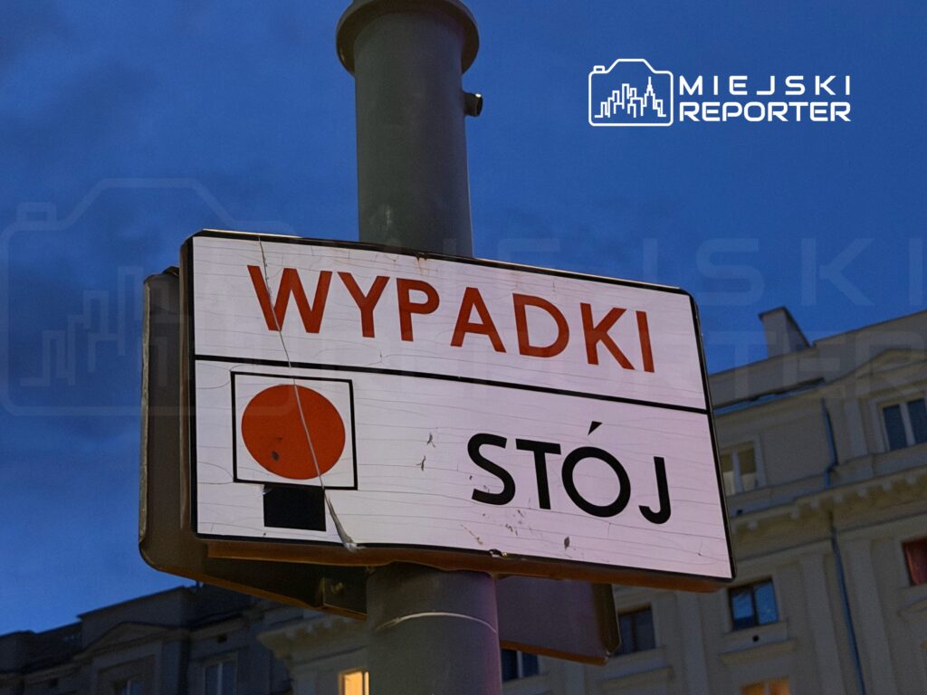 Znak drogowy z napisem "WYPADKI" i "STÓJ" w otoczeniu miejskich budynków.