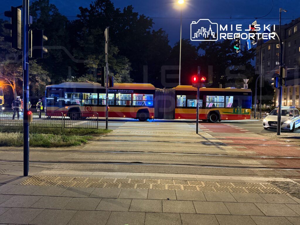 Autobus elektryczny zatrzymany na przejściu dla pieszych, obok policjanci i sygnalizacja świetlna w nocy.