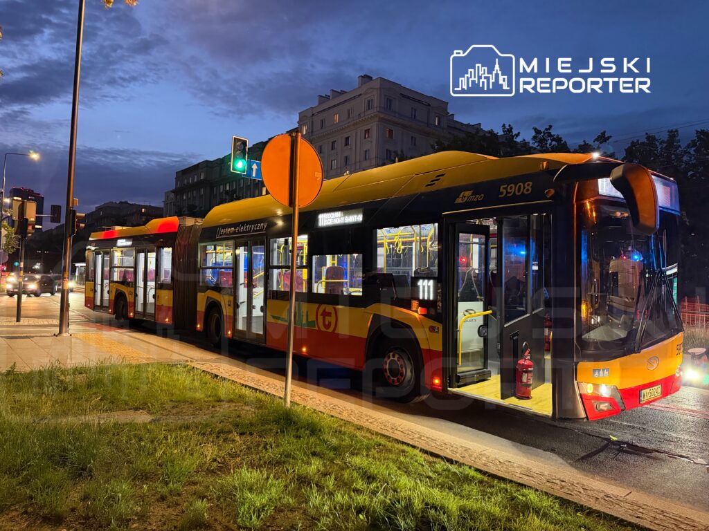 Elektryczny autobus stoi na przystanku, w tle widoczne są światła uliczne oraz nocne niebo.