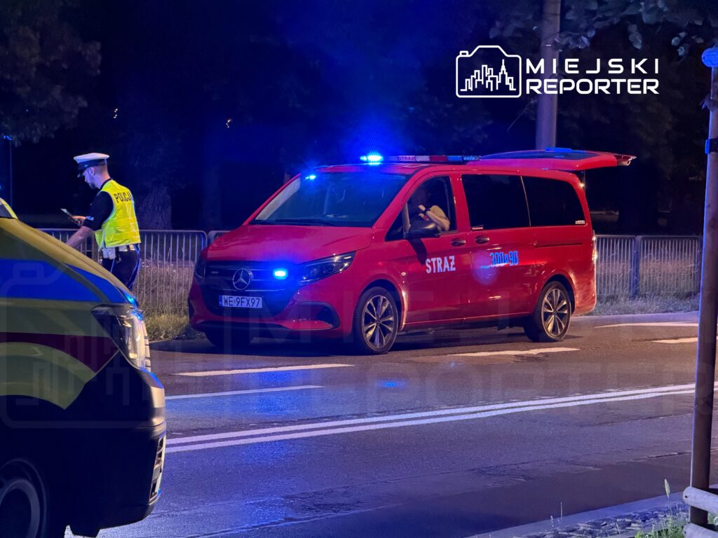 Policjant w kamizelce odblaskowej prowadzi kontrolę drogową obok czerwonego samochodu straży pożarnej z włączonymi sygnałami.