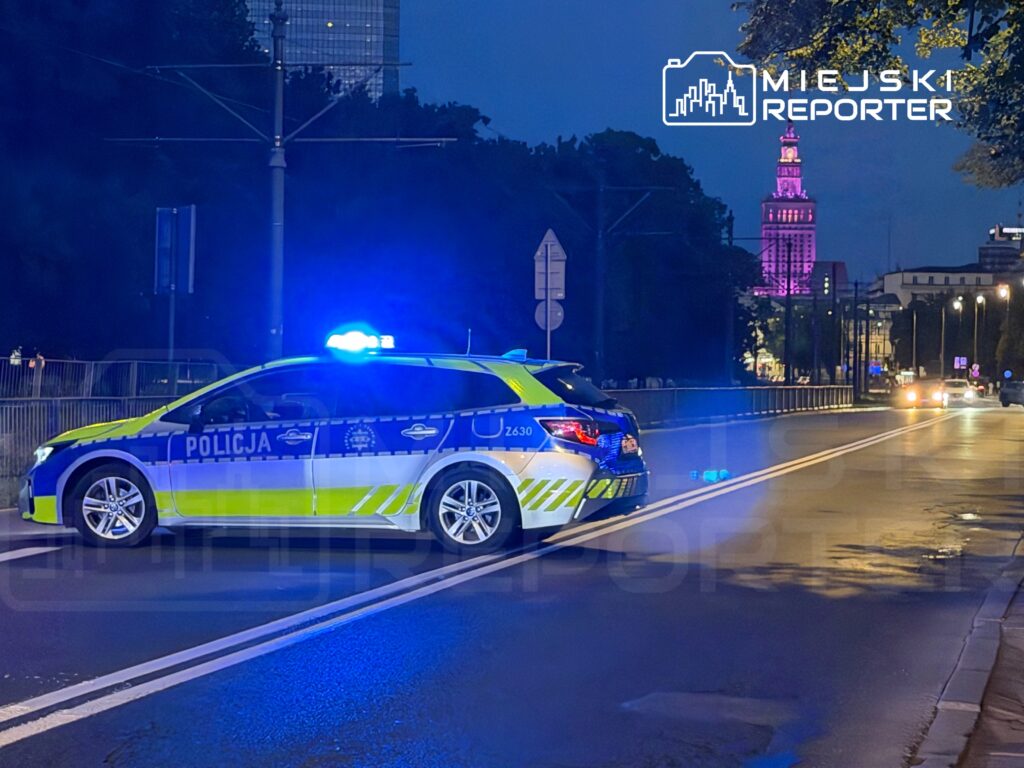 Policyjny radiowóz z włączonymi sygnałami świetlnymi stoi na ulicy nocą, w tle widoczny oświetlony budynek.