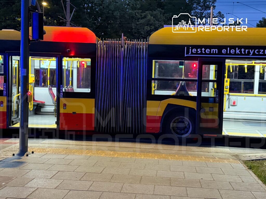 Elektryczny tramwaj z otwartymi drzwiami zatrzymany na przystanku, z widocznymi czerwonymi światłami sygnalizacyjnymi.
