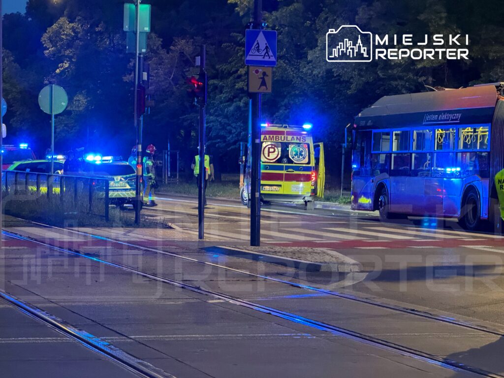 Na ulicy w nocy, zespół ratunkowy i policja przy włączonych sygnałach świetlnych, obok przystanku autobusowego.