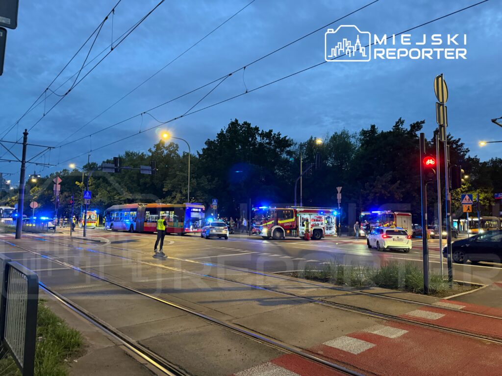 Na skrzyżowaniu ulicy stoją radiowozy, karetki i autobus, a policjant kieruje ruchem w otoczeniu zapalonych sygnalizatorów.