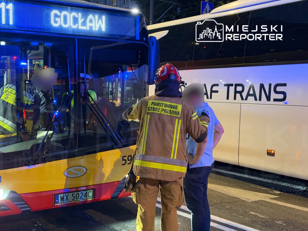 Strażak w mundurze rozmawia z mężczyzną obok przystanku autobusowego, na tle autobusu miejskiego.