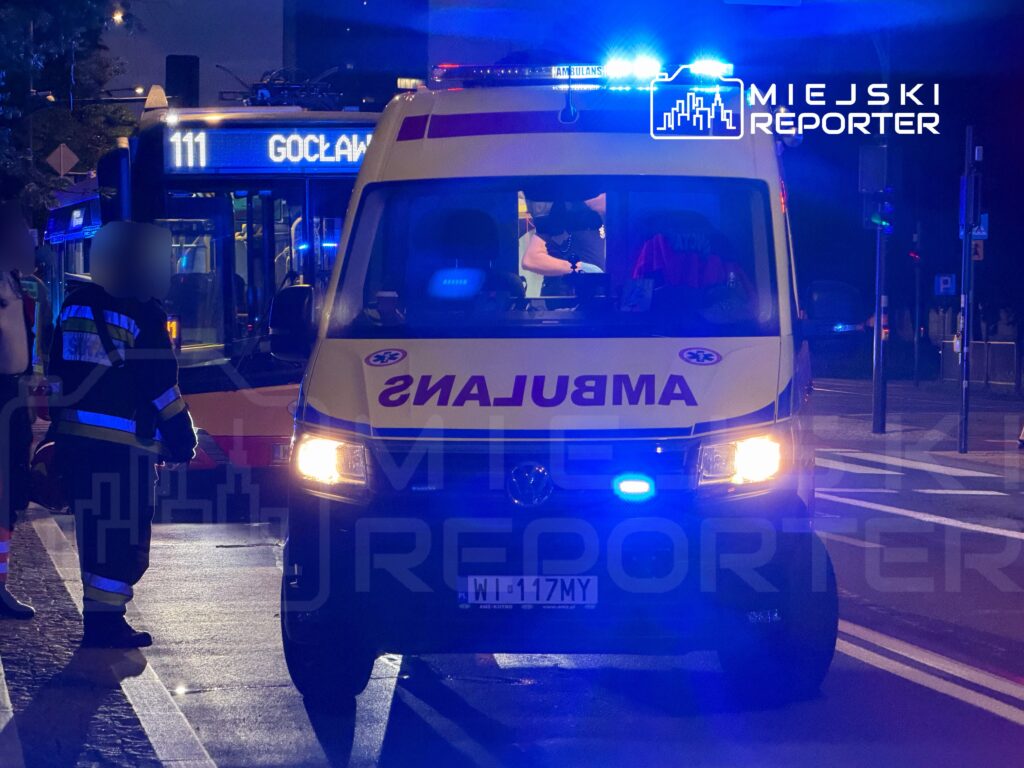 Karetka pogotowia z włączonymi sygnalizatorami świetlnymi zatrzymana obok przystanku autobusowego, przy którym stoi ratownik.