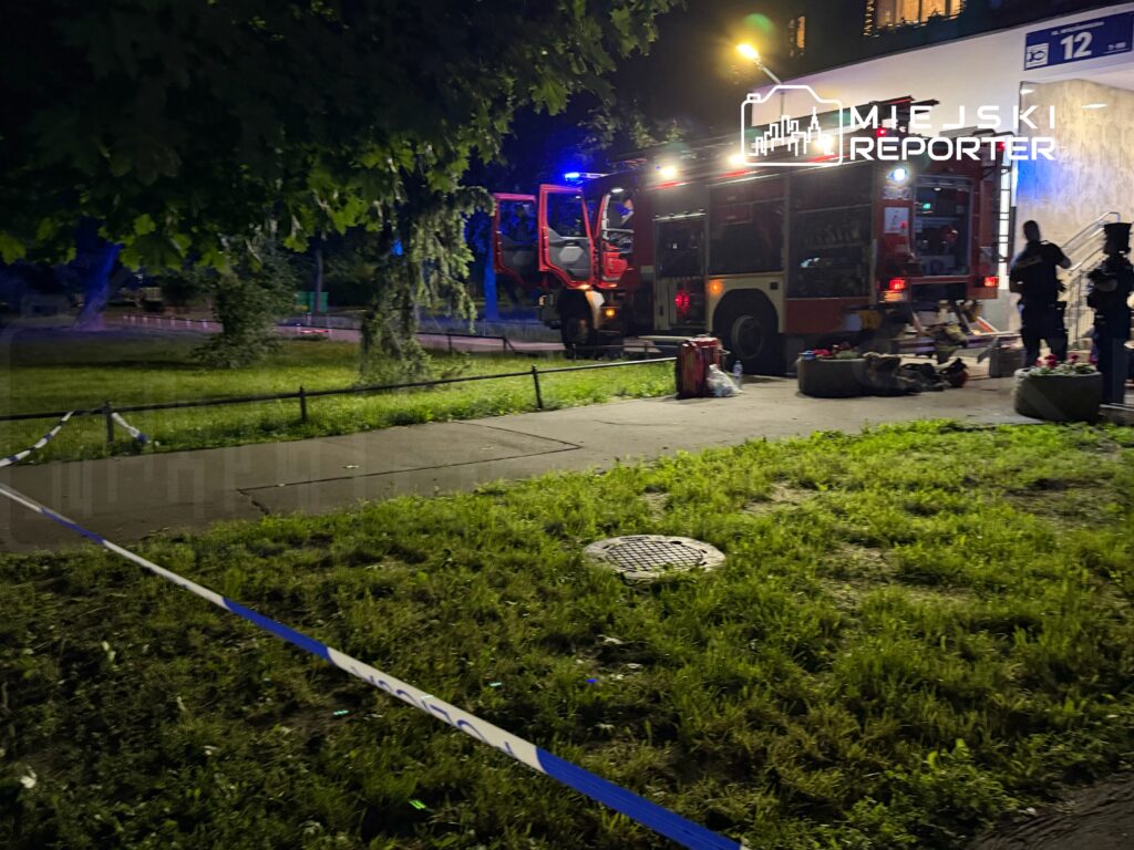 Strażacy w wozie strażackim interweniują przy budynku, otoczeni przez policyjne taśmy zabezpieczające.