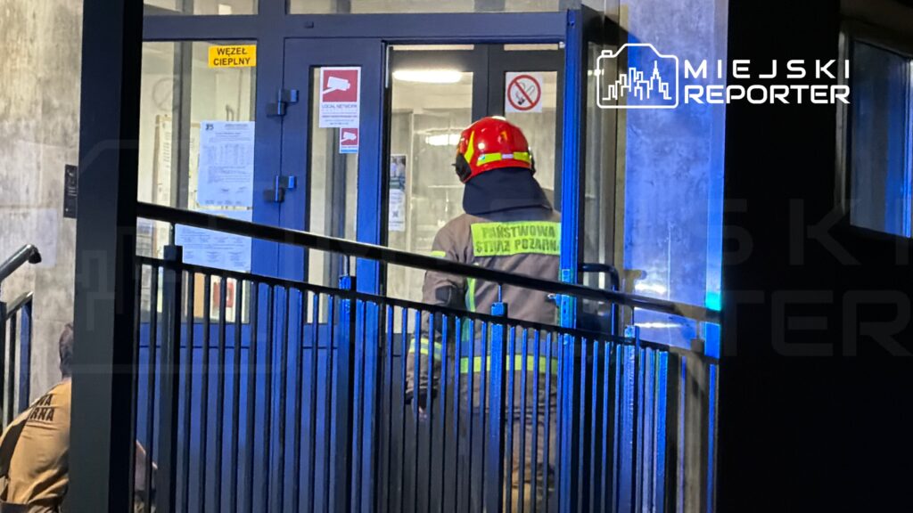 Strażak w hełmie i mundurze wchodzi do budynku oznaczonego jako "WĘZEŁ CIEPLNY", w tle widać niebieskie światło.