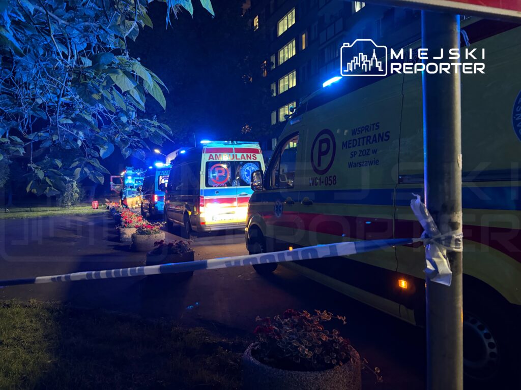 Zespół ambulansów z niebieskimi światłami zaparkowany w nocy na ulicy, w tle budynki mieszkalne.