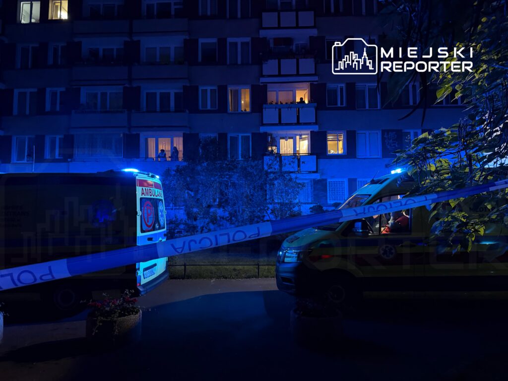 Nocna interwencja służb ratunkowych w pobliżu bloku mieszkalnego, z niebieskimi światłami w karetce i policyjnym taśmą zabezpieczającą.