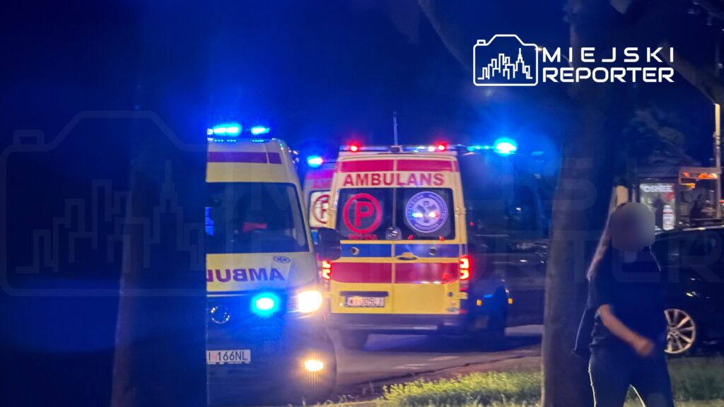 Dwa ambulanse z włączonymi sygnałami świetlnymi stoją na ulicy w nocy, obok przechodząca kobieta.