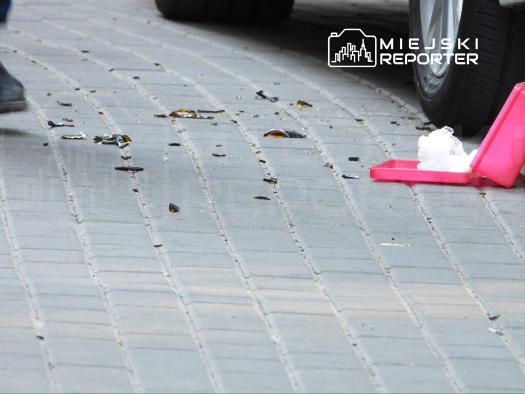 Na chodniku leżą rozbite kawałki szkła oraz różowa plastikowa pudełko z białą gąbką.
