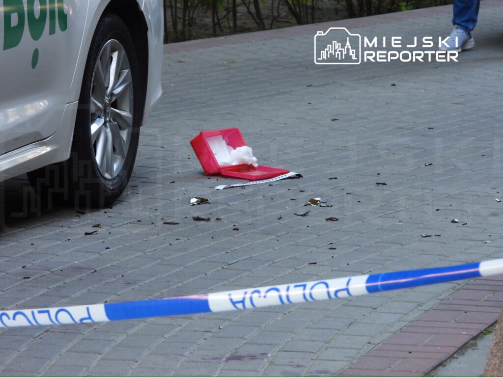 Na chodniku leży czerwona teczka i kawałki szkła, obok zaparkowany samochód z napisem „Dostawa”.