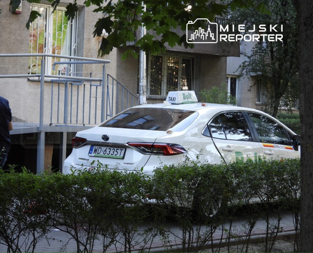 Biały samochód osobowy marki Toyota z oznaczeniem "Taxi Bolt" zaparkowany w zielonym otoczeniu przy budynku mieszkalnym.