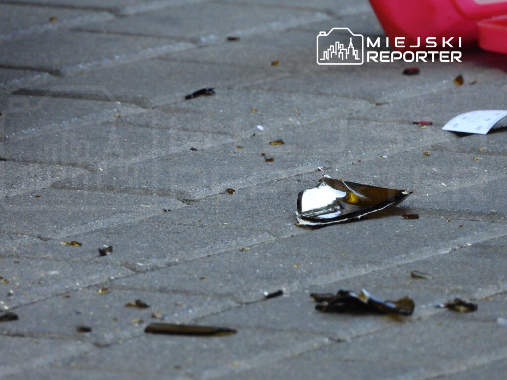 Na chodniku leżą fragmenty rozbitego szkła i drobne śmieci, w tle widoczna jest różowa plastikowa torba.