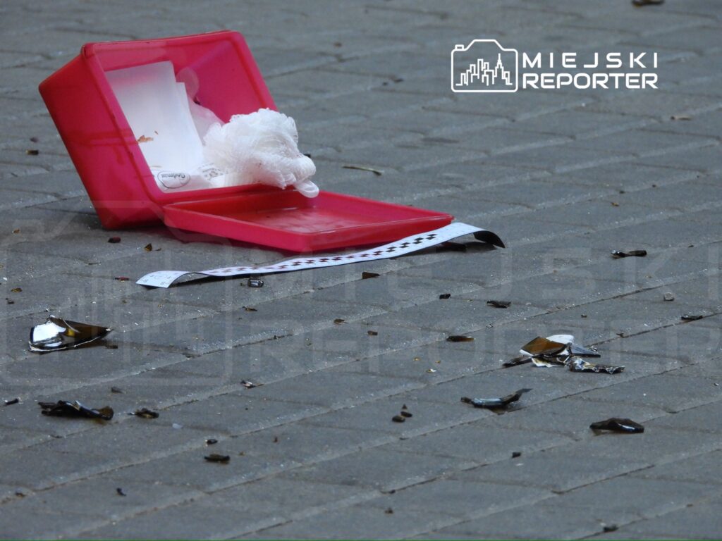 Czerwony pojemnik leży na chodniku, obok niego porozrzucane kawałki szkła i białe fragmenty materiału.