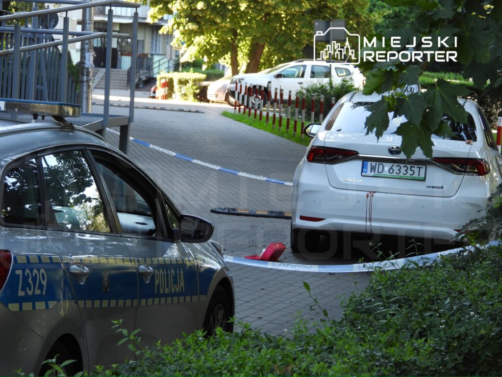 Policyjny radiowóz zaparkowany obok białego samochodu osobowego, w otoczeniu taśm zabezpieczających na chodniku.